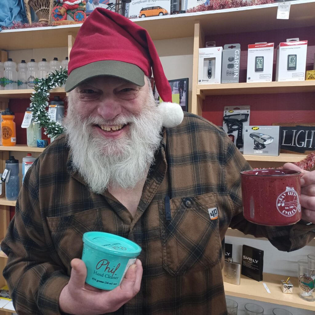 Older man with beard wearing a santa hat baseball cap and holding a mug and tub of soap.