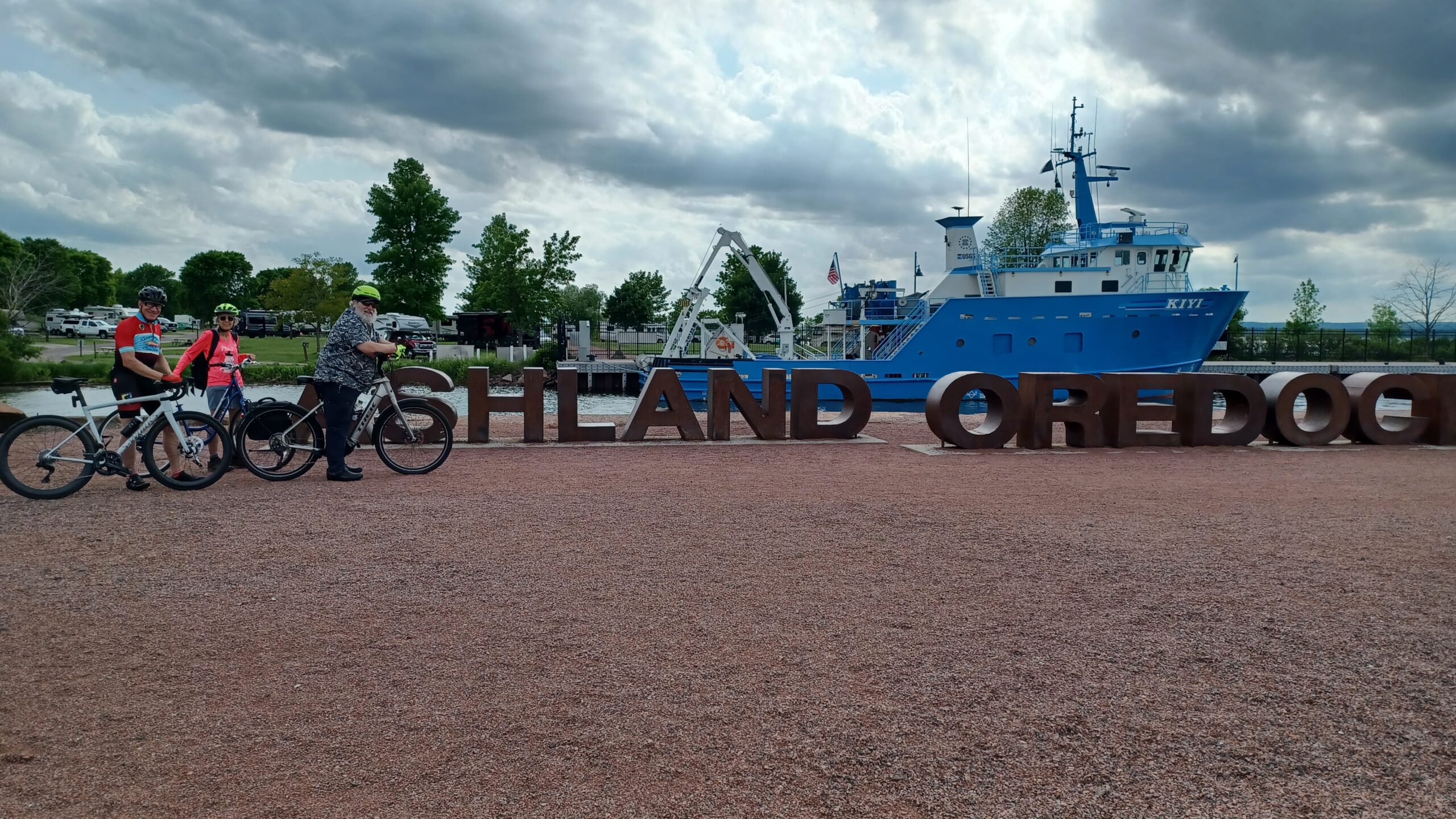 Group of cyclists standing next to statue which reads "Ashland Oredock" with blue boat in the background.